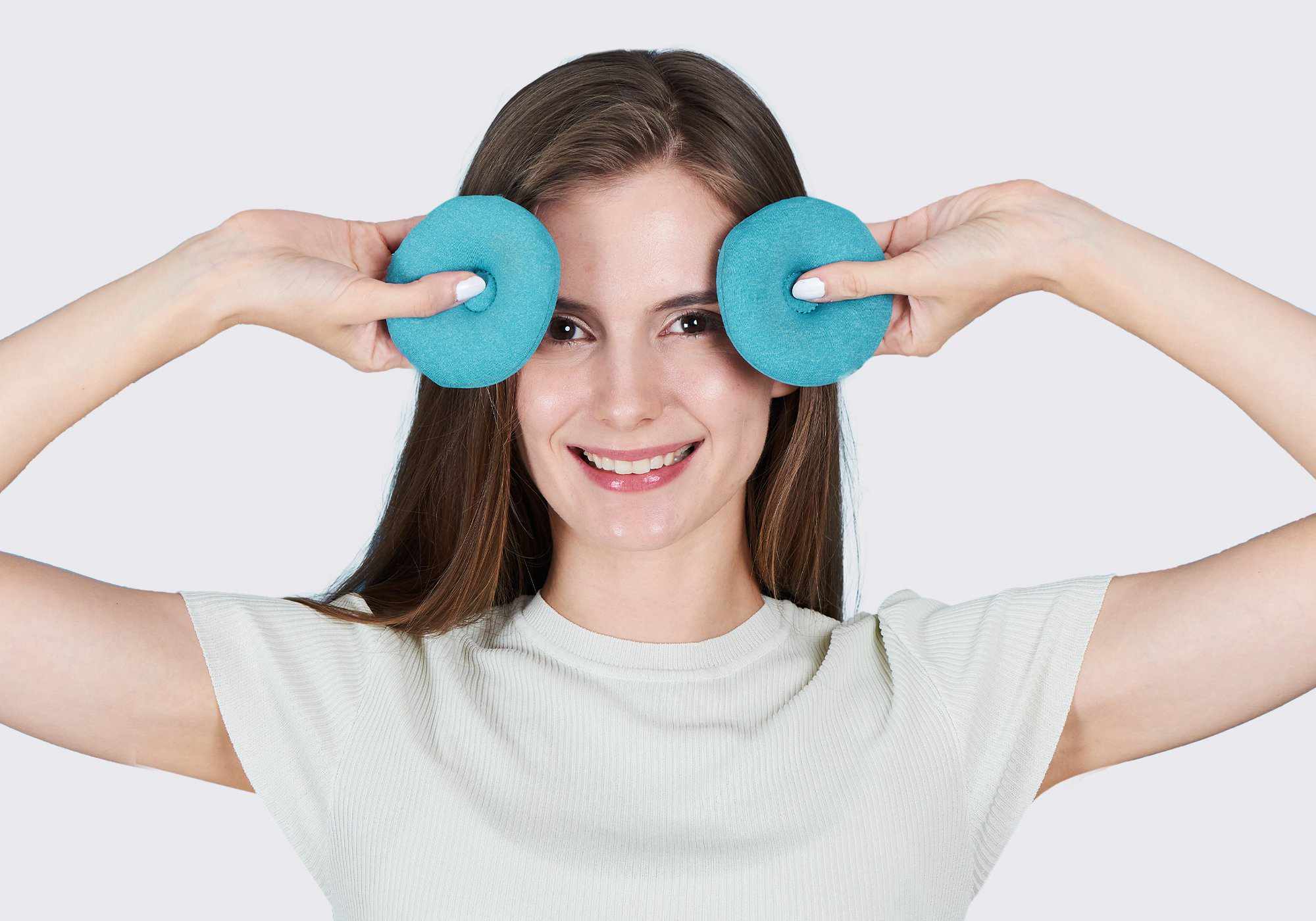 A smiling woman holding two round, blue-teal fabric eye cups over her eyes against a plain light gray background.
