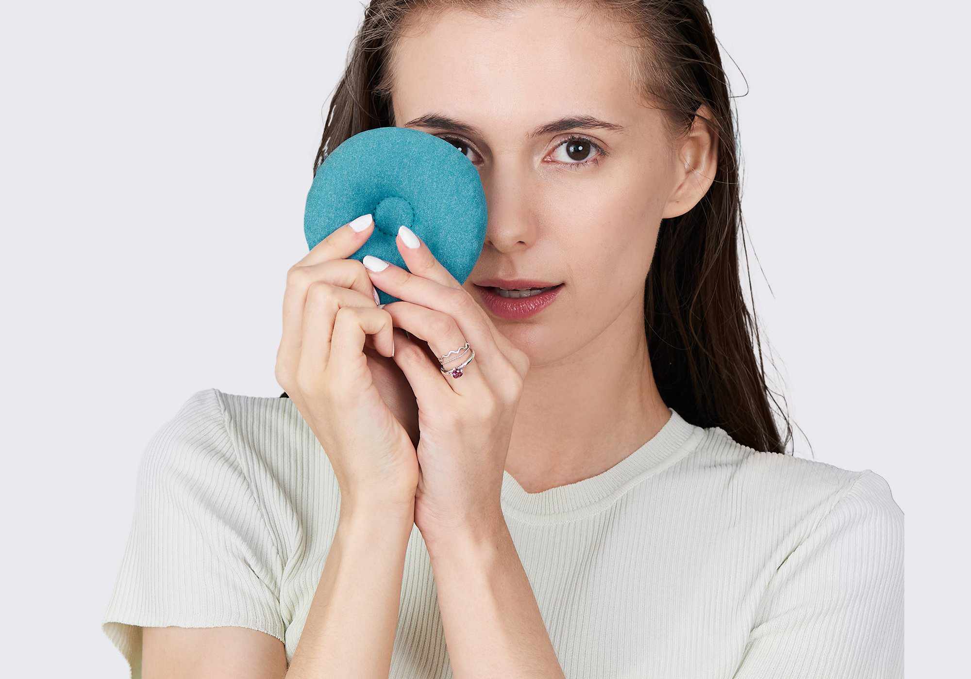 A woman posing while holding a circular, blue-teal fabric eye cup over one eye against a light gray background.