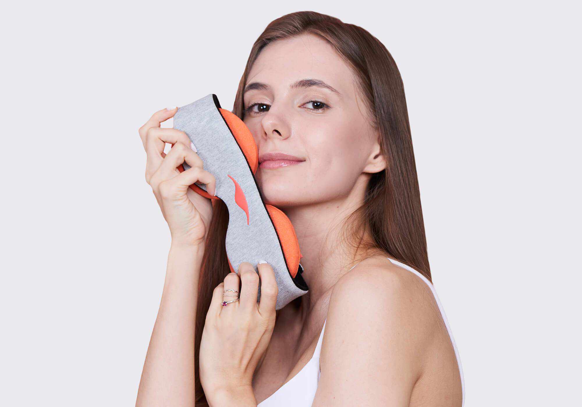 A close-up lifestyle portrait of a person with straight brown hair, holding the Manta STEAM Sleep Mask near their face against a plain light background. They are wearing a white tank top and a silver ring on their finger, presenting a gentle smile as they highlight the contrast between the gray heathered band and the round orange eye cups.