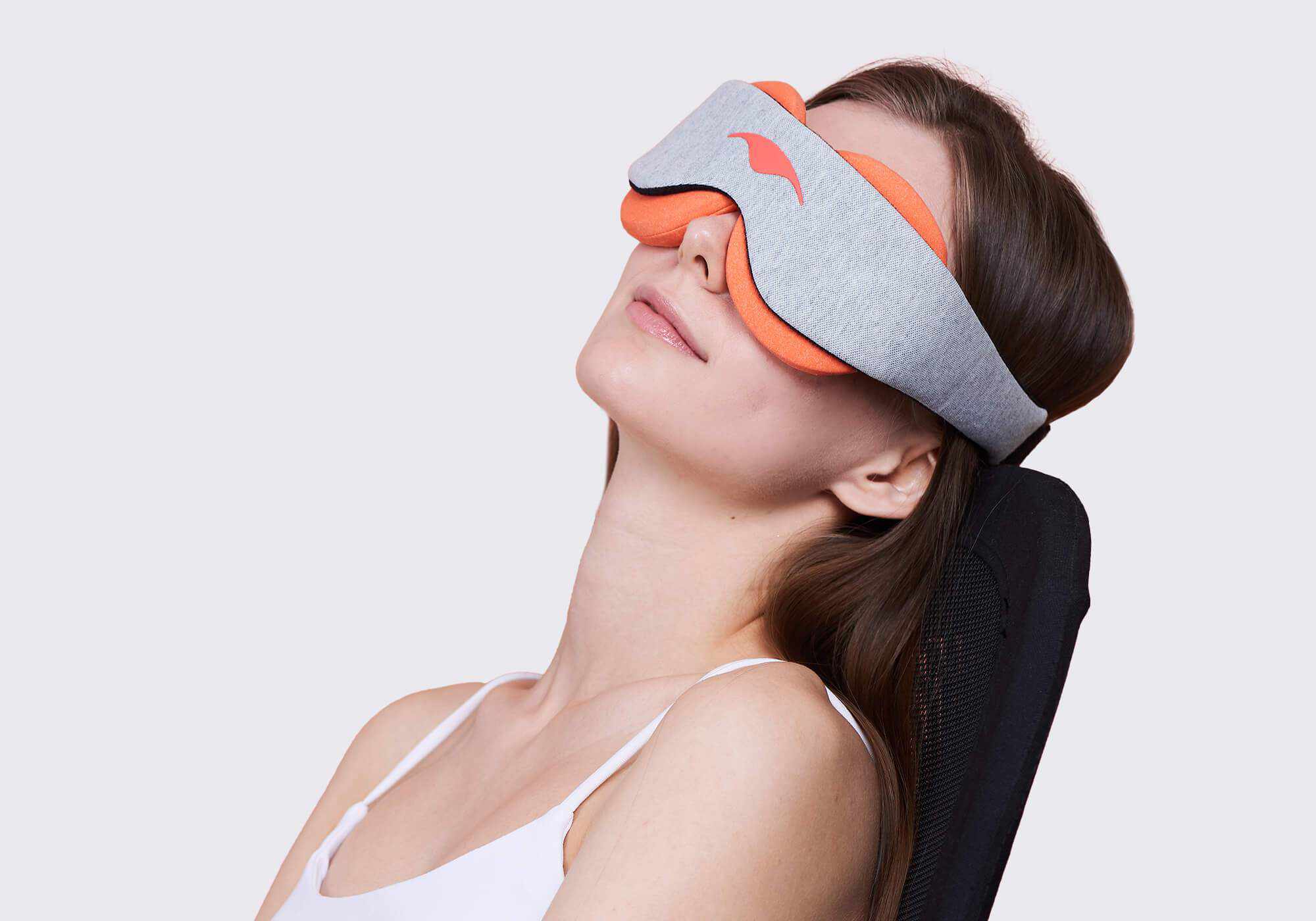 A portrait shot from a lower angle showing a person with long brown hair leaning back in a black chair, wearing the Manta STEAM Sleep Mask over their eyes. The mask features a light gray heathered front band and thick, circular terracotta-orange eye cups that rest securely against the contours of their face.