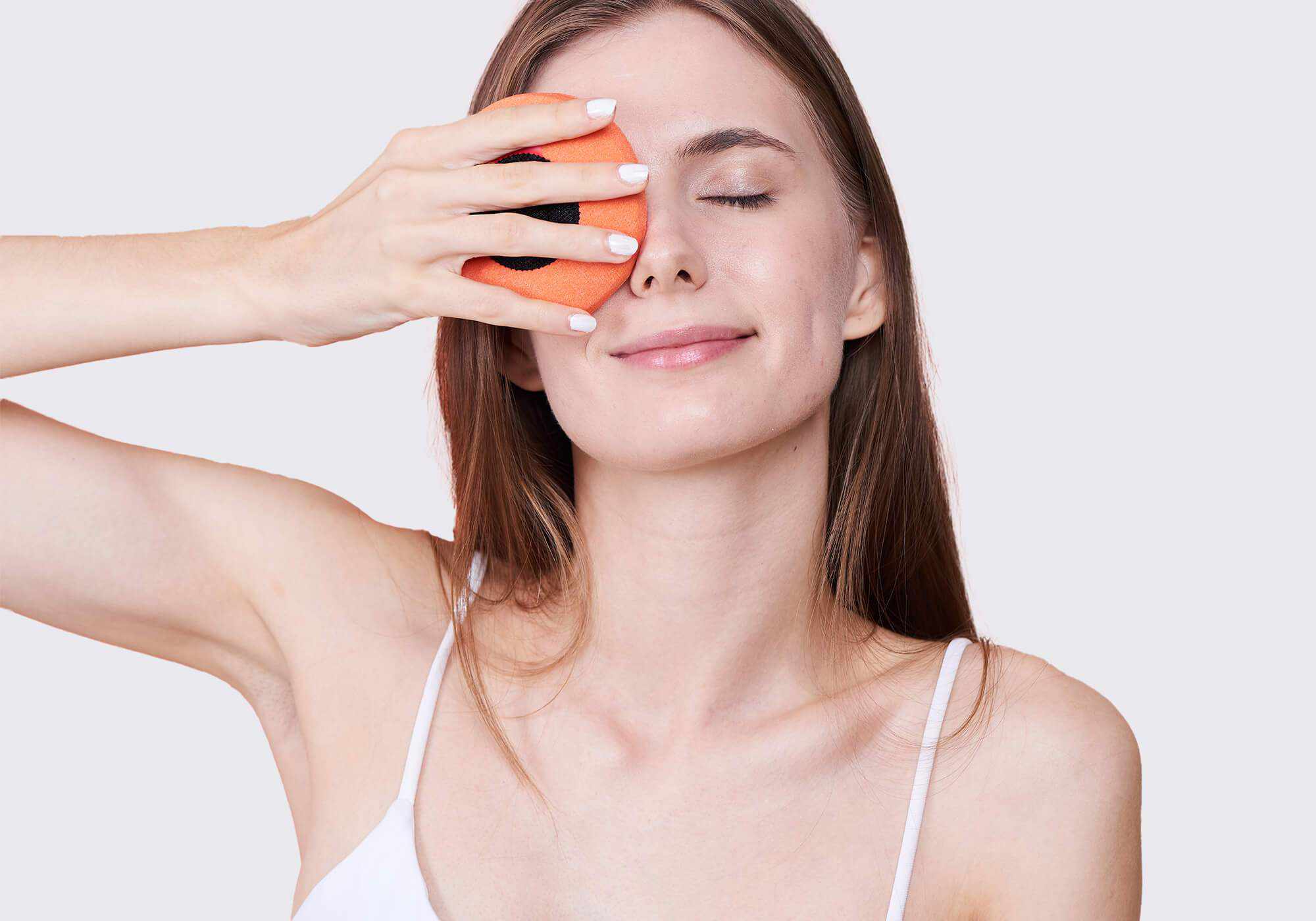 A woman with eyes closed smiling gently while holding a circular orange fabric eye cup over one eye against a light gray background.