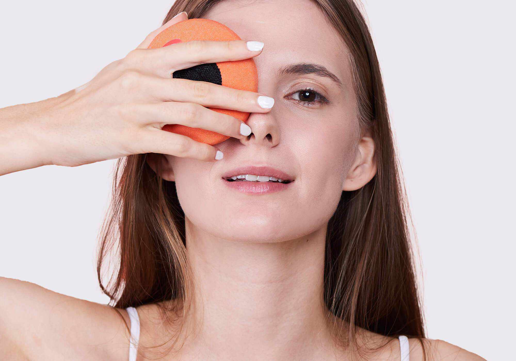A woman with white nail polish smiling while holding a circular orange fabric eye cup over one eye against a light gray background.