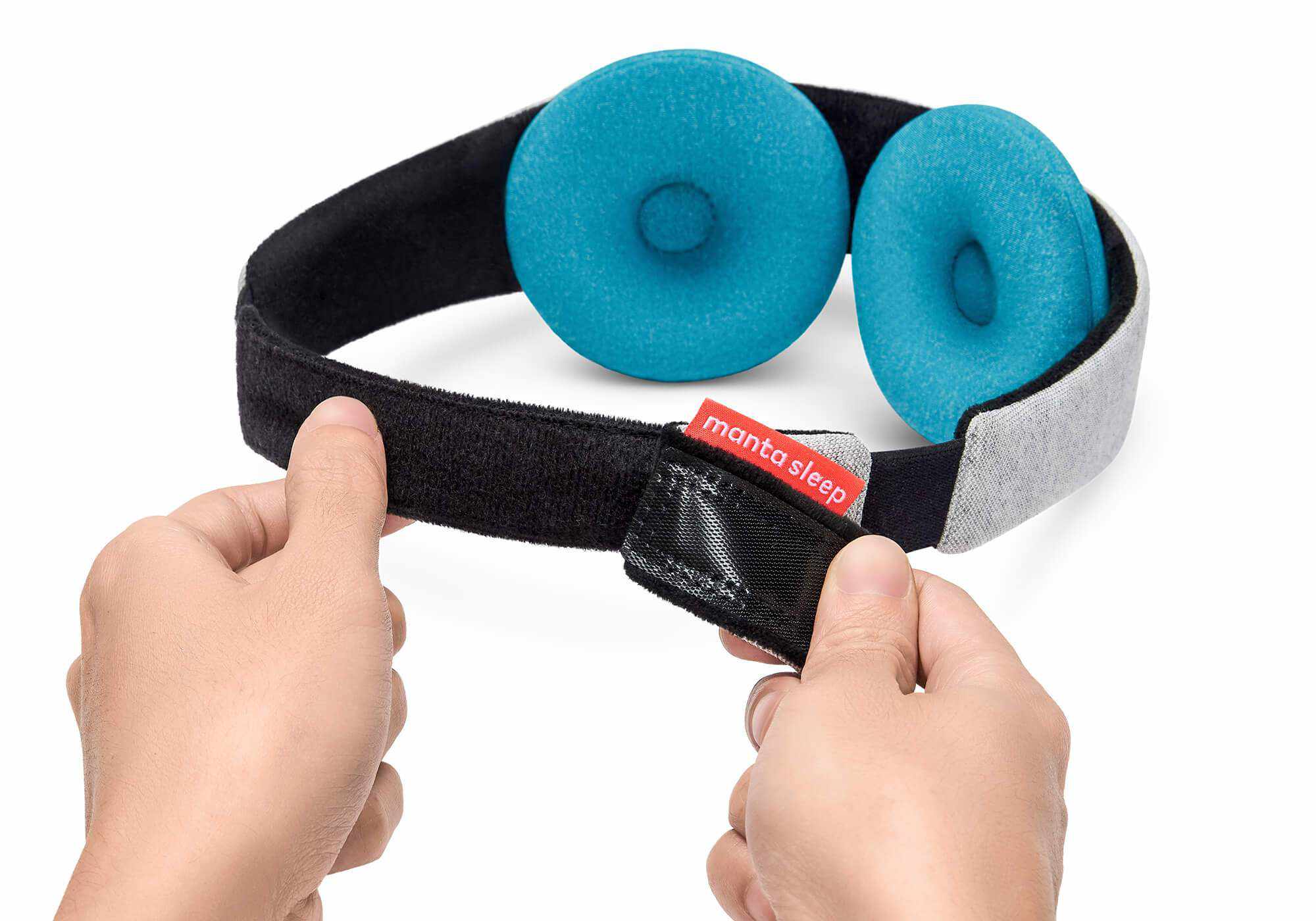 Hands adjusting the black hook-and-loop back closure of a grey Manta COOL Sleep Mask strap fitted with round blue-teal eye cups.