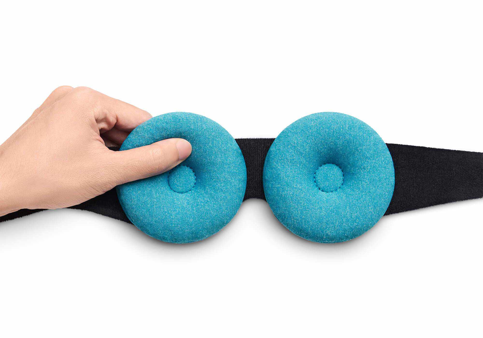 A hand pressing into a circular blue-teal Manta COOL Eye Cup attached to a black loop and hook on a head strap, isolated on a white background.