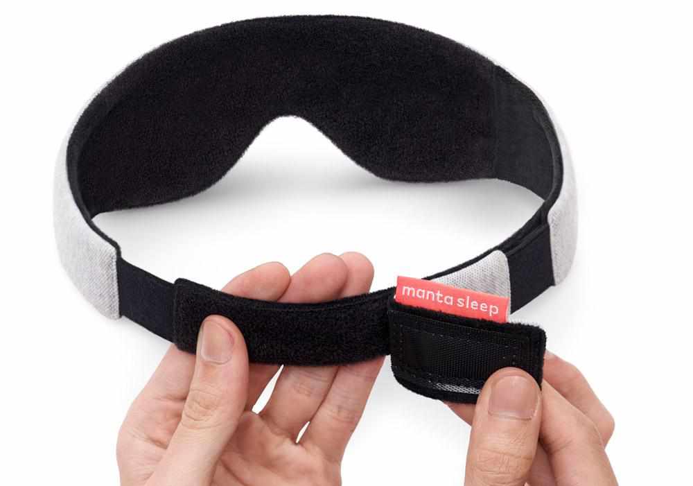 Hands adjusting the black hook-and-loop closure at the back of the grey sleep mask strap, showing the red Manta Sleep tag.