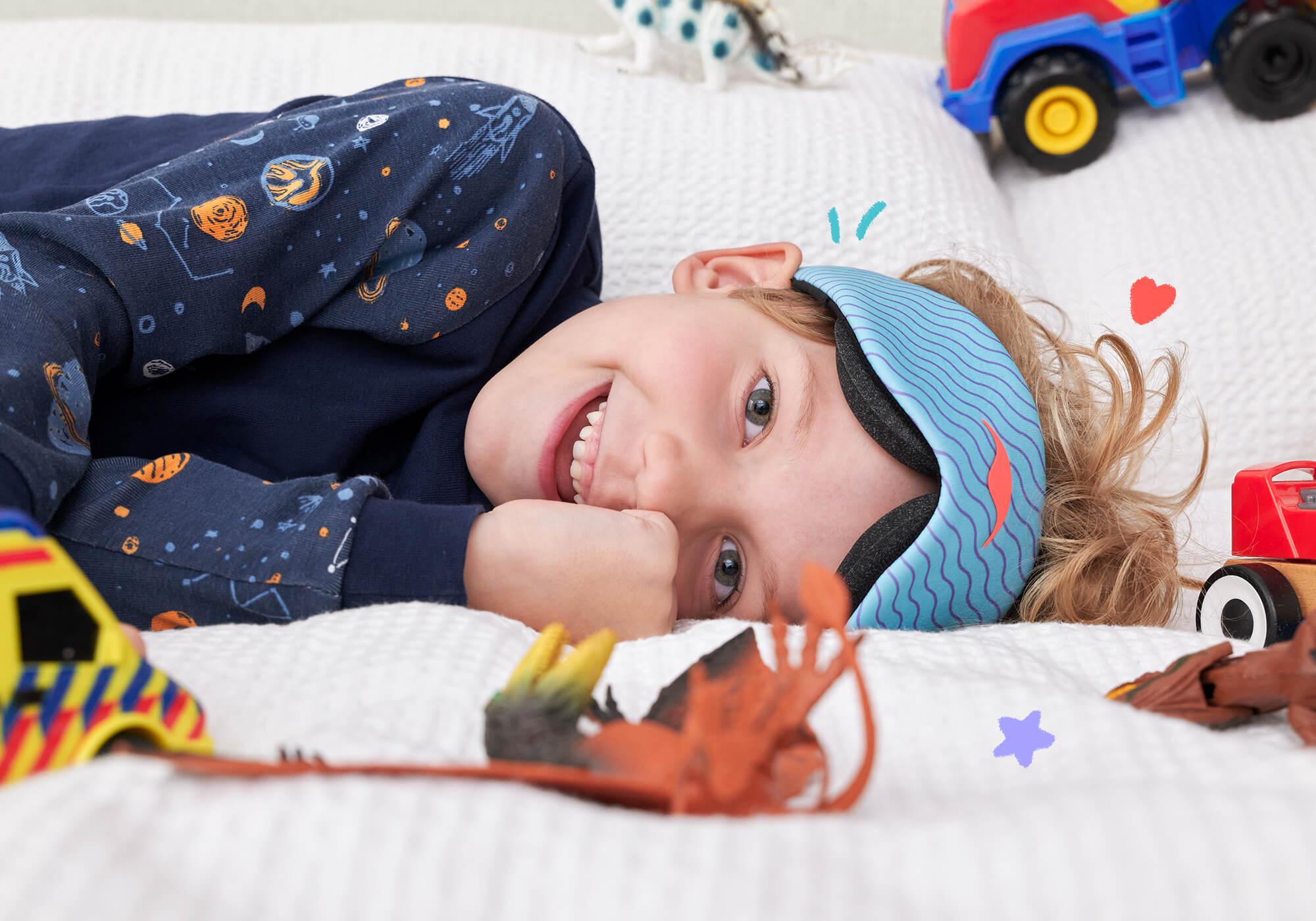 A child wearing space-patterned pajamas lies flat on a white bed spread, smiling broadly while wearing a light blue Manta KIDS Sleep Mask with purple wavy lines. Various toy cars and action figures are scattered around them on the mattress.