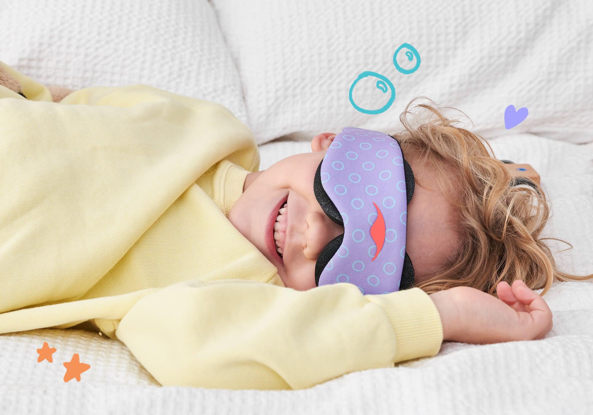 A close-up lifestyle shot of a smiling child with wavy hair lying on a white textured bedspread, wearing a yellow sweatshirt and the purple patterned Manta KIDS Sleep Mask over their eyes. The image features whimsical hand-drawn digital doodles of bubbles, stars, and a small heart floating around the child.