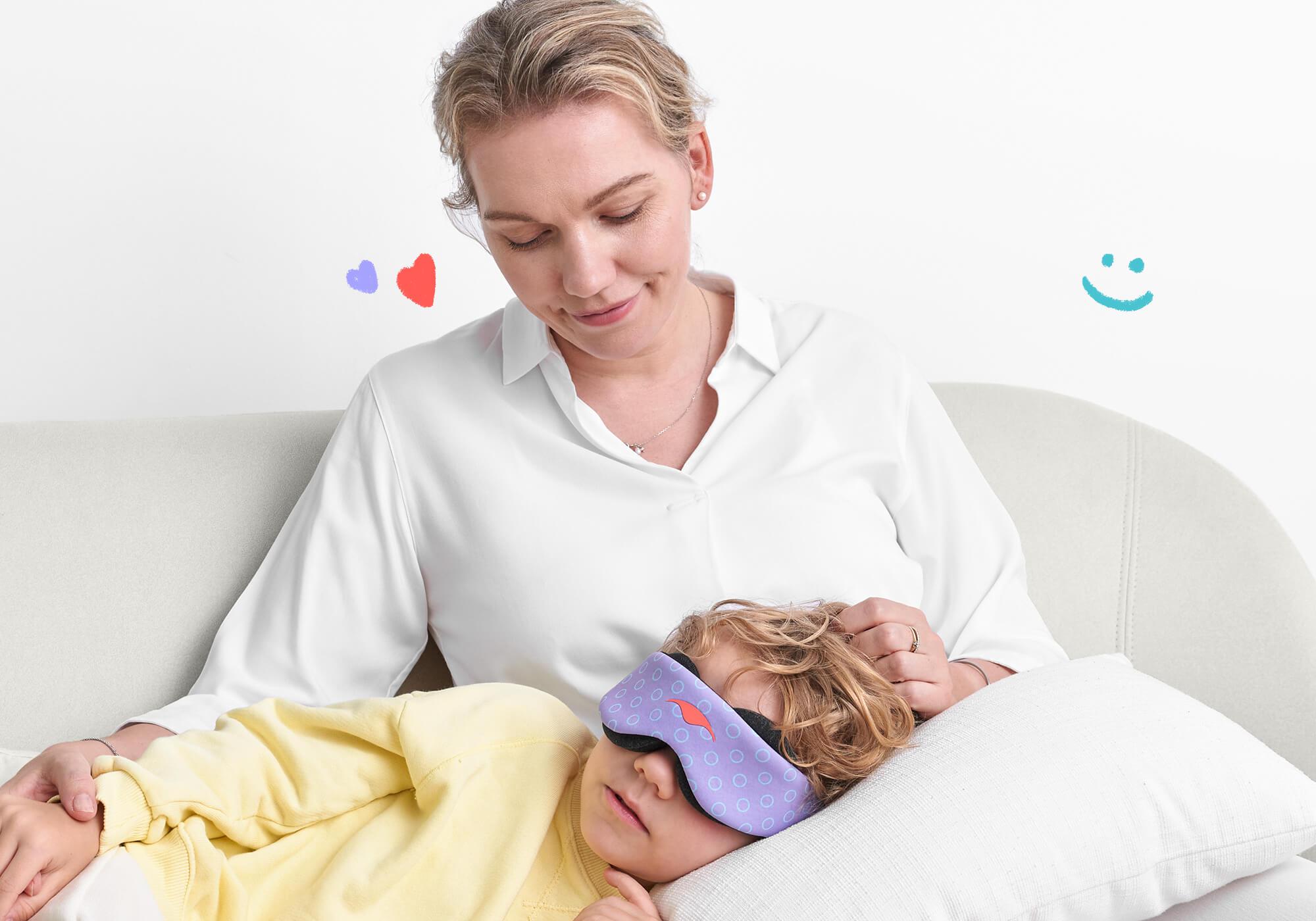 An adult wearing a white collared shirt sits on a sofa, looking down warmly at a child with wavy hair who is resting their head on a white pillow in their lap. The child is wearing a purple patterned Manta KIDS Sleep Mask. Small hand-drawn digital doodles of hearts and a smiley face are scattered in the background.