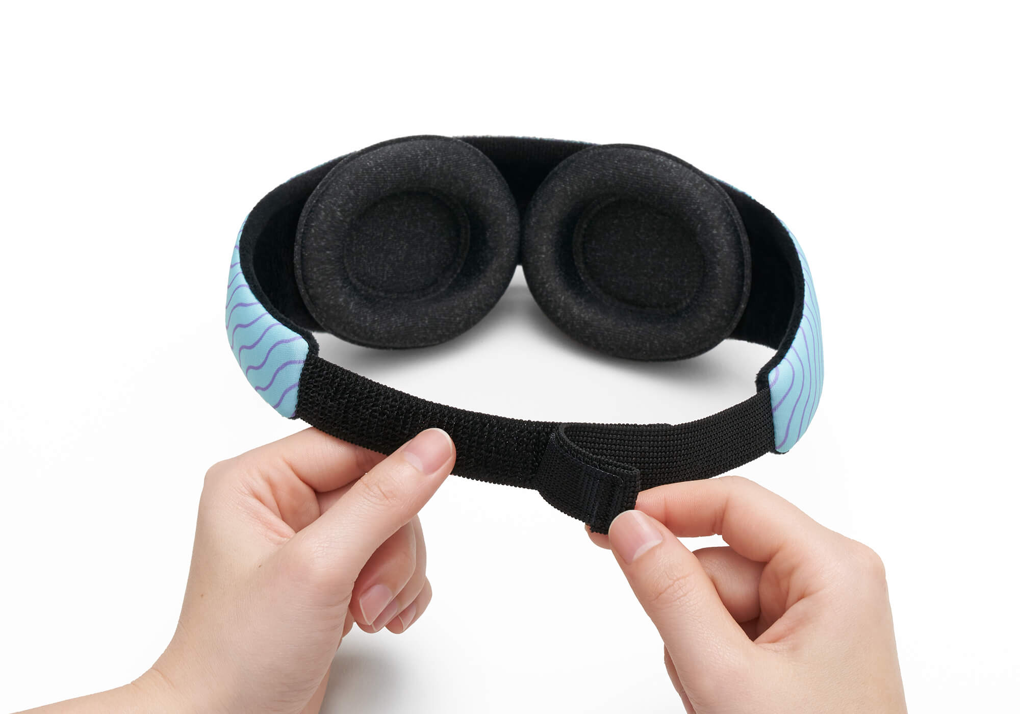 A close-up view of two hands fastening the micro-velcro closure area at the back of a light blue Manta KIDS Sleep Mask. The interior reveals two modular, contoured black fabric eye cups attached to a plush black backing, positioned against a stark white background.