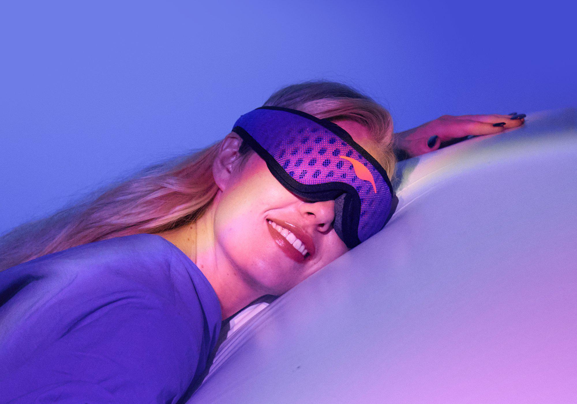 Close-up of a woman smiling while resting her head, wearing a purple and blue textured Manta Sleep Mask PRO under neon lighting.