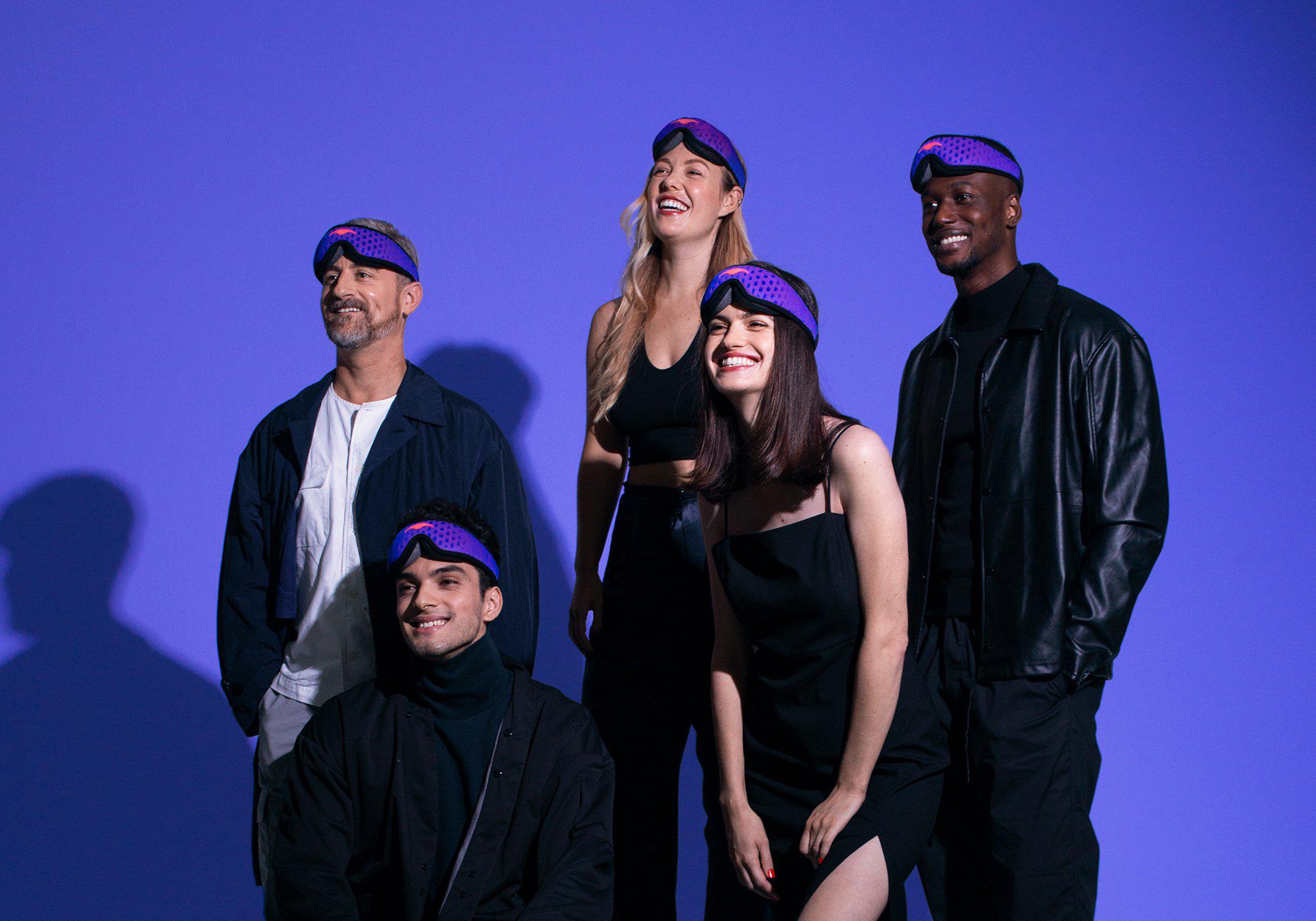 A group of five people smiling and looking upward while wearing purple Manta Sleep Mask PROs on their foreheads under blue lighting.