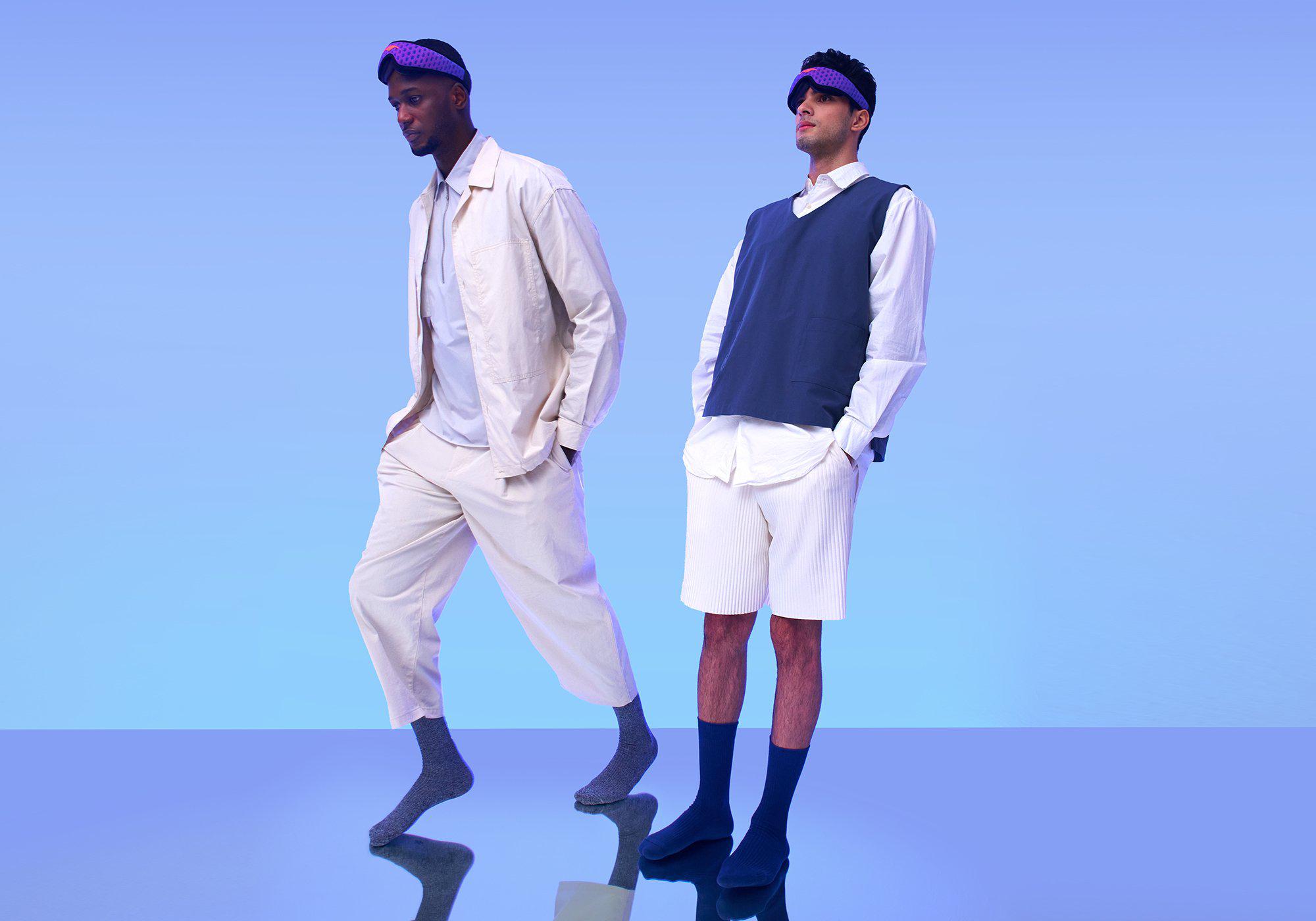 Two men modeling light-colored, casual outfits and wearing purple Manta Sleep Mask PROs on their foreheads against a light blue backdrop.