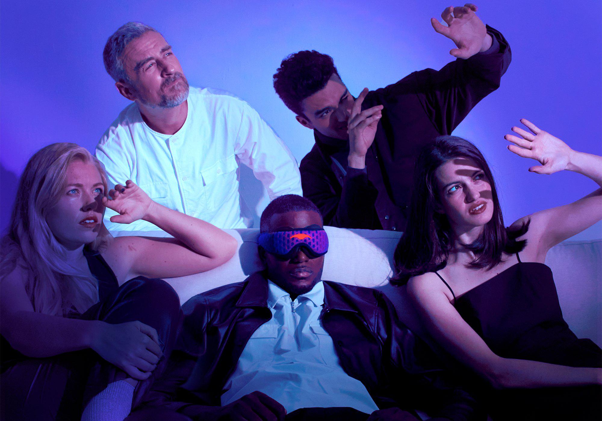 Five people on a couch under harsh blue light shields their eyes from glare, except one man wearing a black-out Manta PRO Sleep Mask.