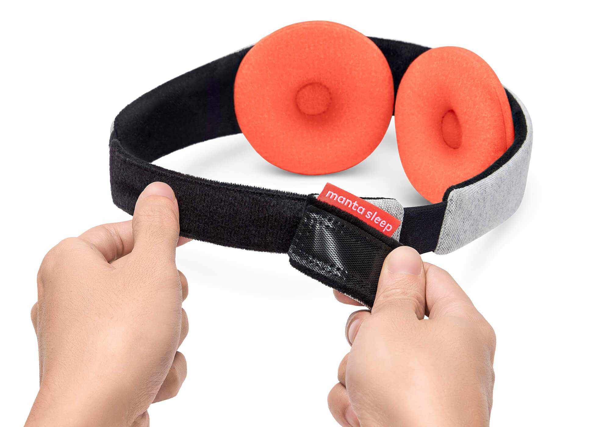 A close-up view of two hands separating the hook-and-loop closure at the back of the Manta STEAM Sleep Mask's elastic band. A small red fabric tag with the white text "Manta Sleep" is stitched near the closure, while the light gray heathered outer strap and thick round orange eye cups are visible in the background against a white surface.