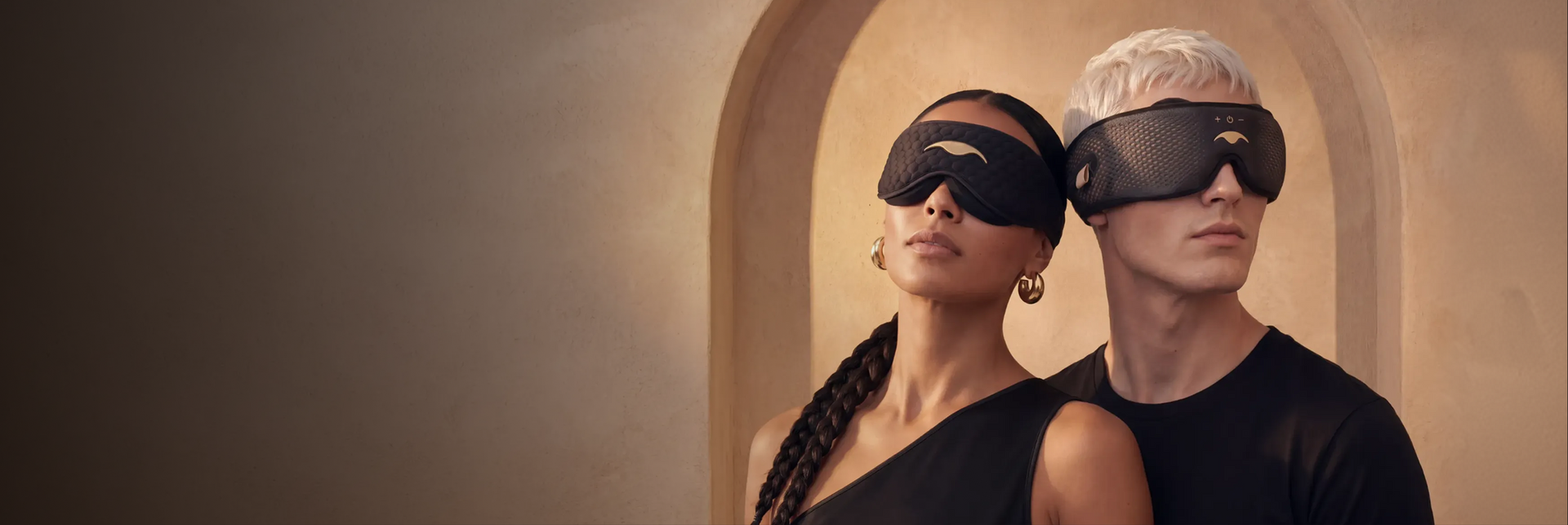 Two people next to each other, one wears Manta SILK Sleep Mask and other wears Manta SOUND Sleep Mask, both masks are in a black-gold colorway.