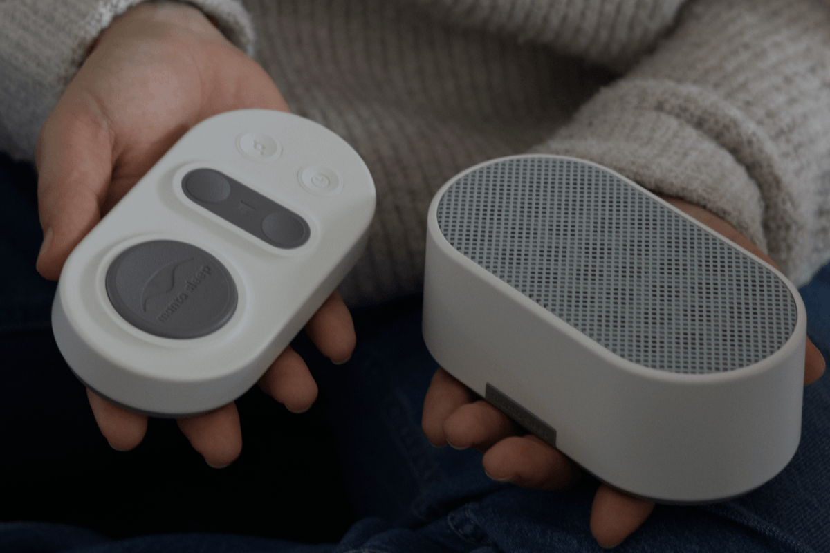 A close-up of hands holding Stone White Manta White Noise Machine speaker and remote pad against a dark background.