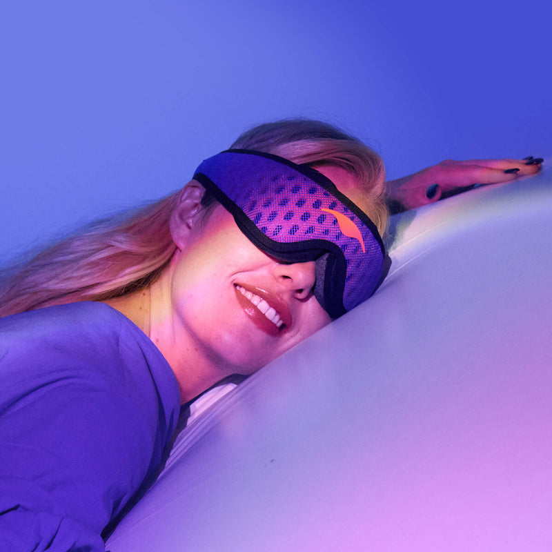 Close-up of a woman smiling while resting her head, wearing a purple and blue textured Manta Sleep Mask PRO under neon lighting.