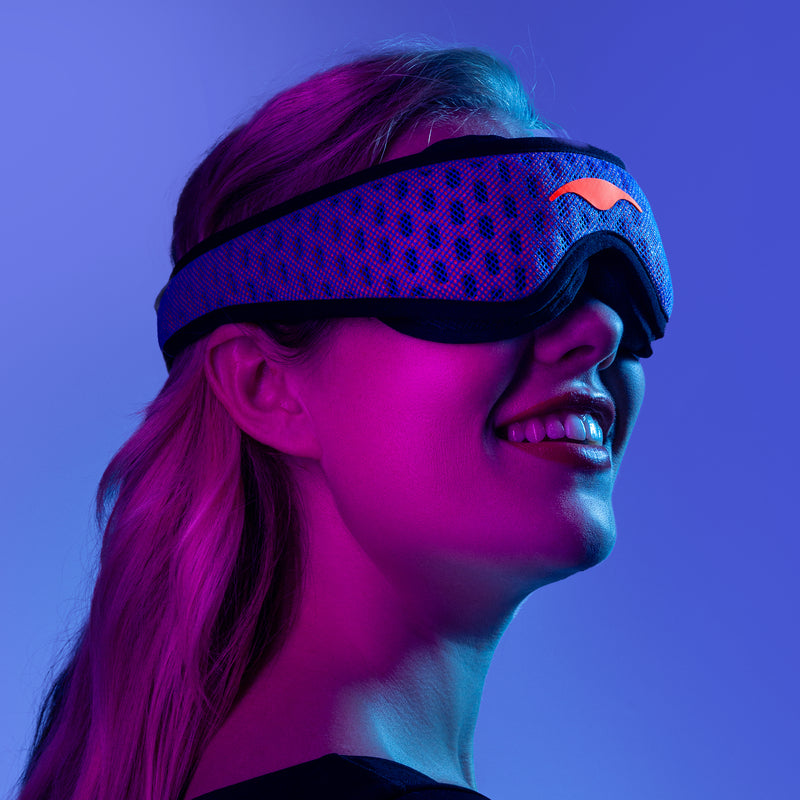 Smiling blonde person wearing a blue and purple Manta PRO Sleep Mask, in a dark setting, illuminated with purple light on a blue background.