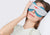A woman resting her head on a white pillow while wearing a grey Manta COOL Sleep Mask fitted with blue-teal eye cups.