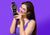 A smiling woman with colorful round aroma patches stuck on her face holds up the interior side of a Manta Sleep Mask, against a solid purple background.