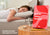 A red Manta Sleep Journal on a wooden table next to a candle and a glass of water, with a woman sleeping out of focus in the background.