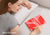 A smiling woman resting her head on a white pillow next to a red Manta Sleep Journal on the bed. Overlay text reads "Quiet your mind".
