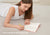 A woman lying on her stomach, writing with a yellow pencil inside an open Manta Sleep Journal. Overlay text reads "Lasts 6 months with daily use".