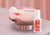 A pink Manta SILK Sleep Mask resting on a metallic silver display stand, set against a soft pink draped fabric background, and a white bottle of Manta Pillow Spray in front of it.
