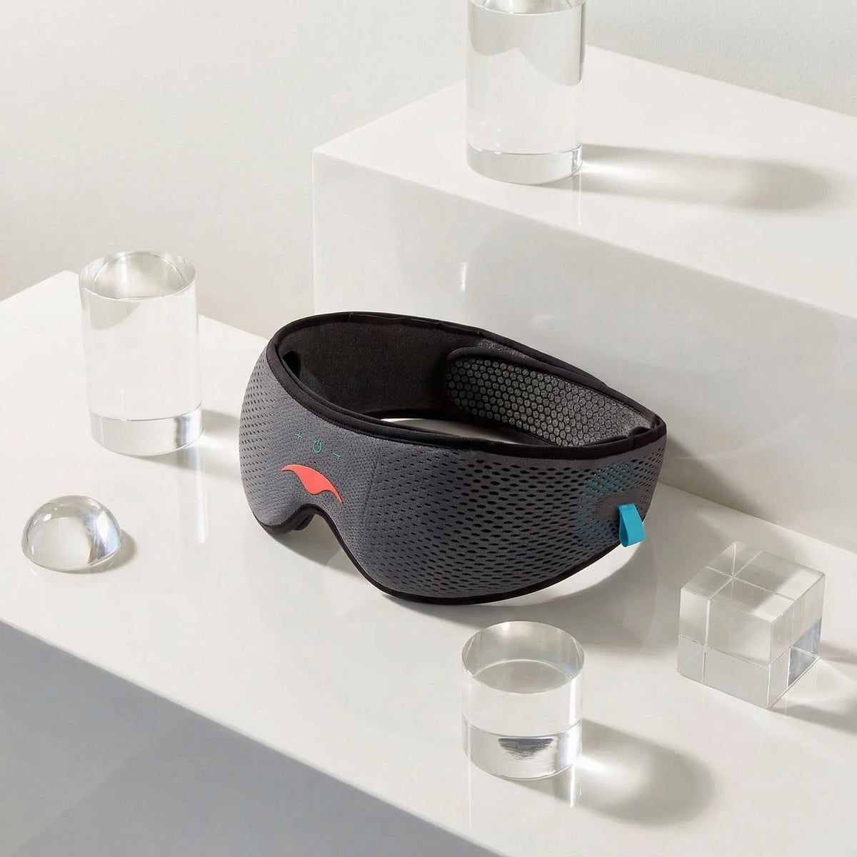 Gray Manta SOUND Sleep Mask on a white surface with transparent glasses and geometric shapes.