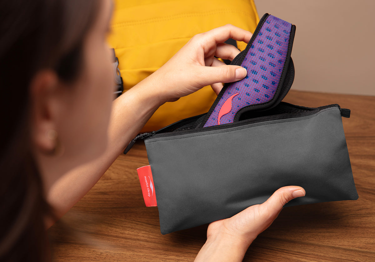 Person opening a black and gray Manta travel pouch to reveal a purple and red Manta Pro Sleep Mask, with a yellow backpack in the background.