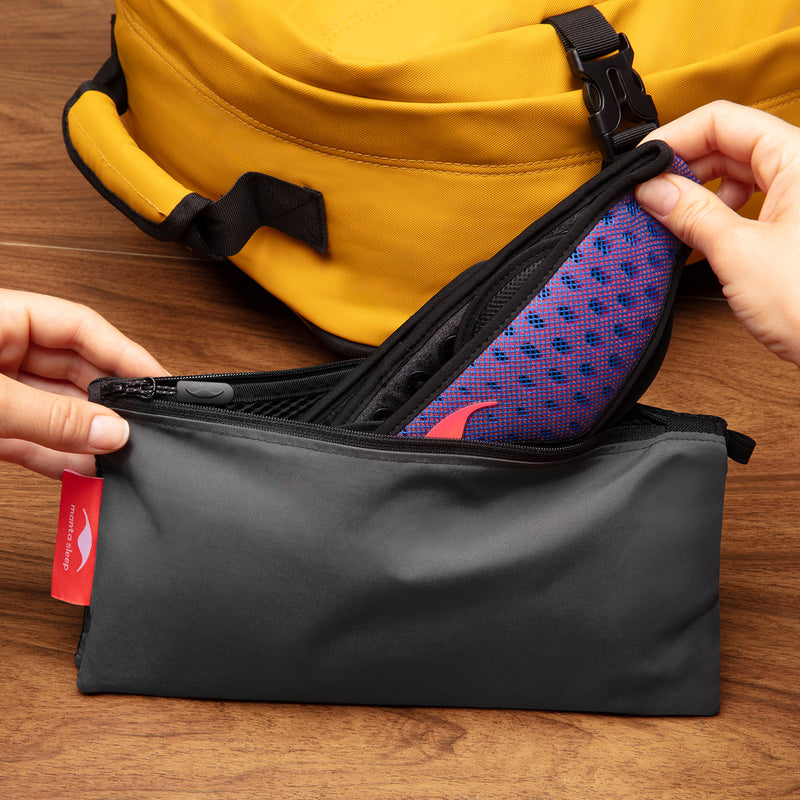 Person opening a black and gray Manta travel pouch to reveal a purple and red Manta Pro Sleep Mask, with a yellow backpack in the background.