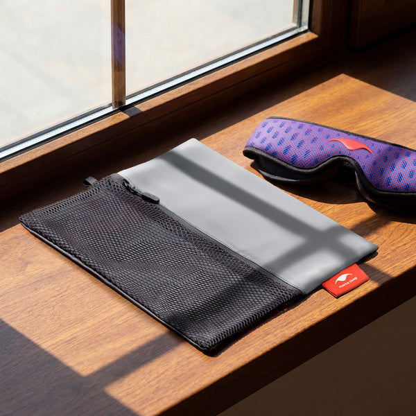 An unfolded grey and black mesh Manta Travel & Laundry Bag resting on a wooden window sill next to a blue and red Manta PRO Sleep Mask in the sunlight.