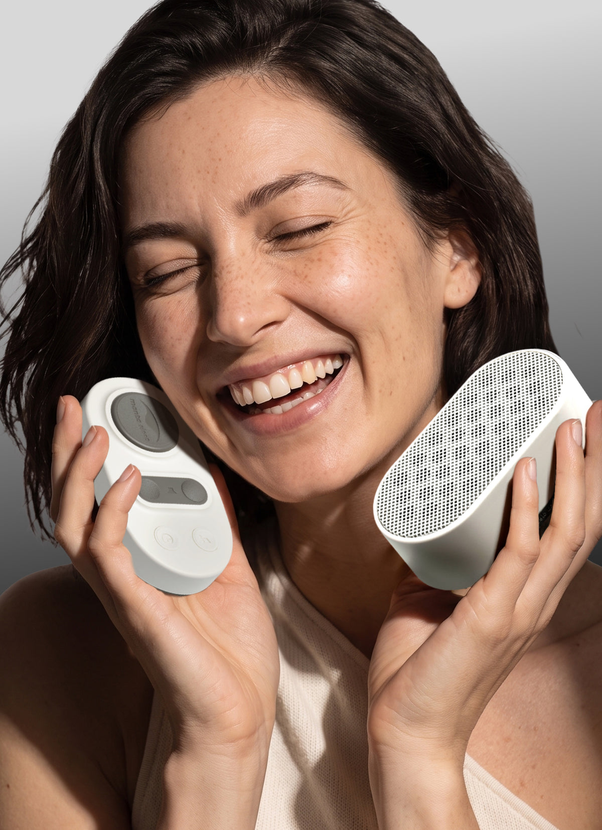 Smiling woman holding the compact Stone White Manta White Noise Machine speaker and wireless remote next to her cheeks.