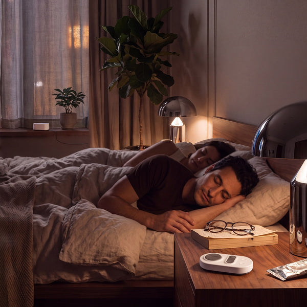 Couple sleeping peacefully in bed with the Stone White Manta White Noise Machine remote resting on a wooden bedside table in foreground.