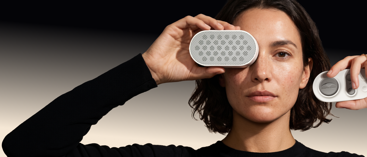 A woman with dark hair holds Manta White Noise Machine in Stone White color over her face against a black background with a white gradient at the bottom.