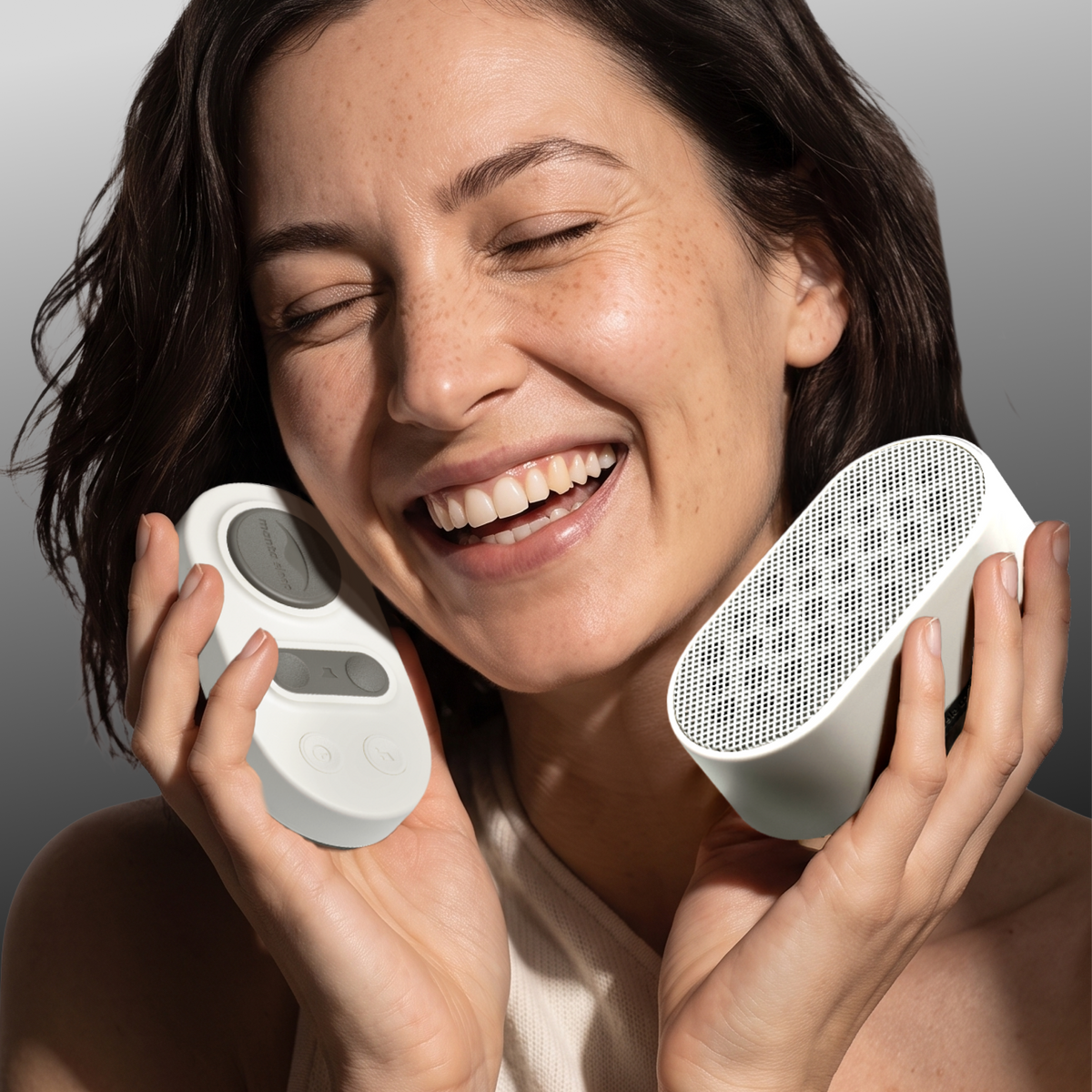 A smiling woman holds Manta White Noise Machine and its remote pad in Stone White color against a clean gray studio background.