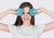 A smiling woman holding two round, blue-teal fabric eye cups over her eyes against a plain light gray background.