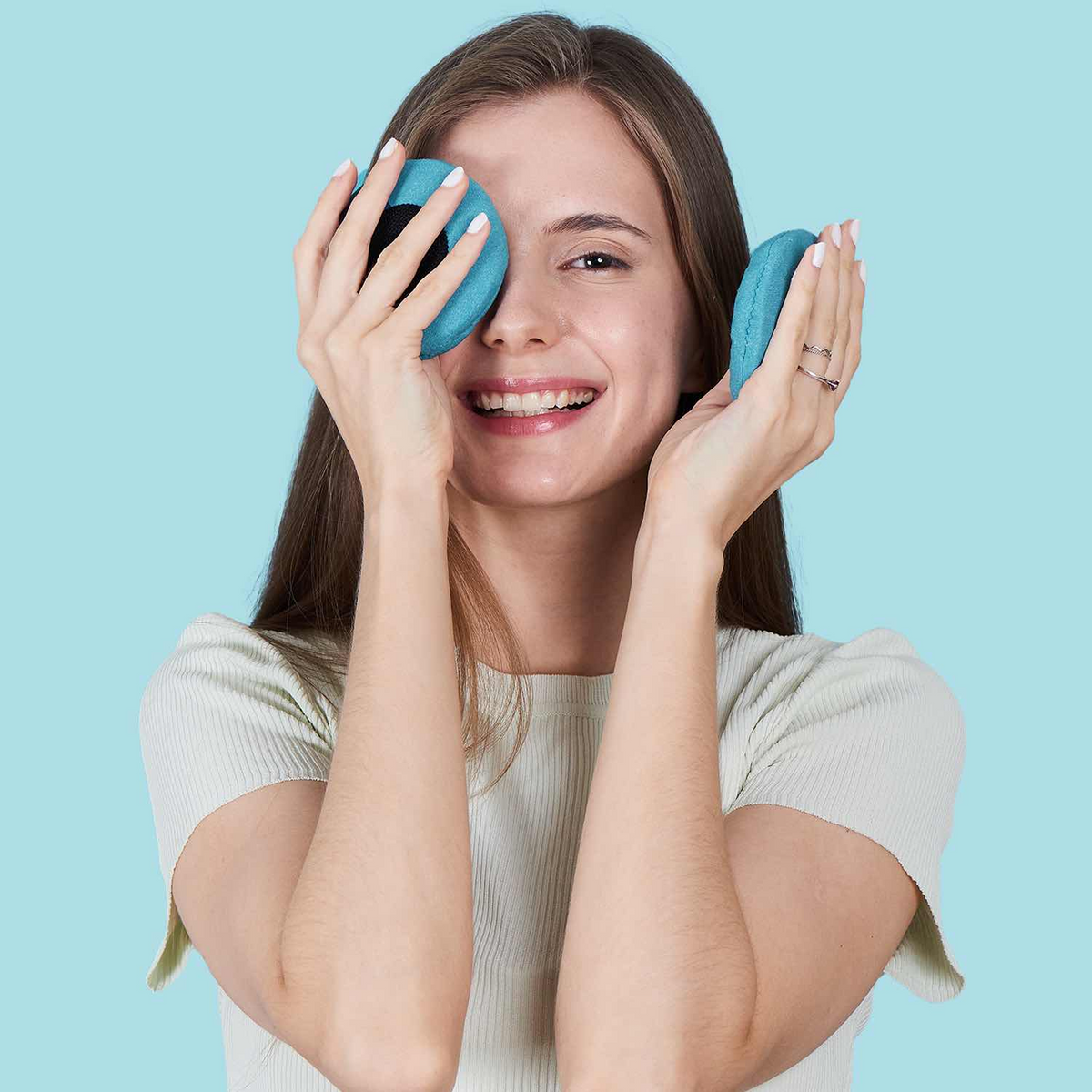 A woman with long brown hair smiles while holding two teal blue Manta COOL Eye Cups, one over each eye, against a solid light blue background.