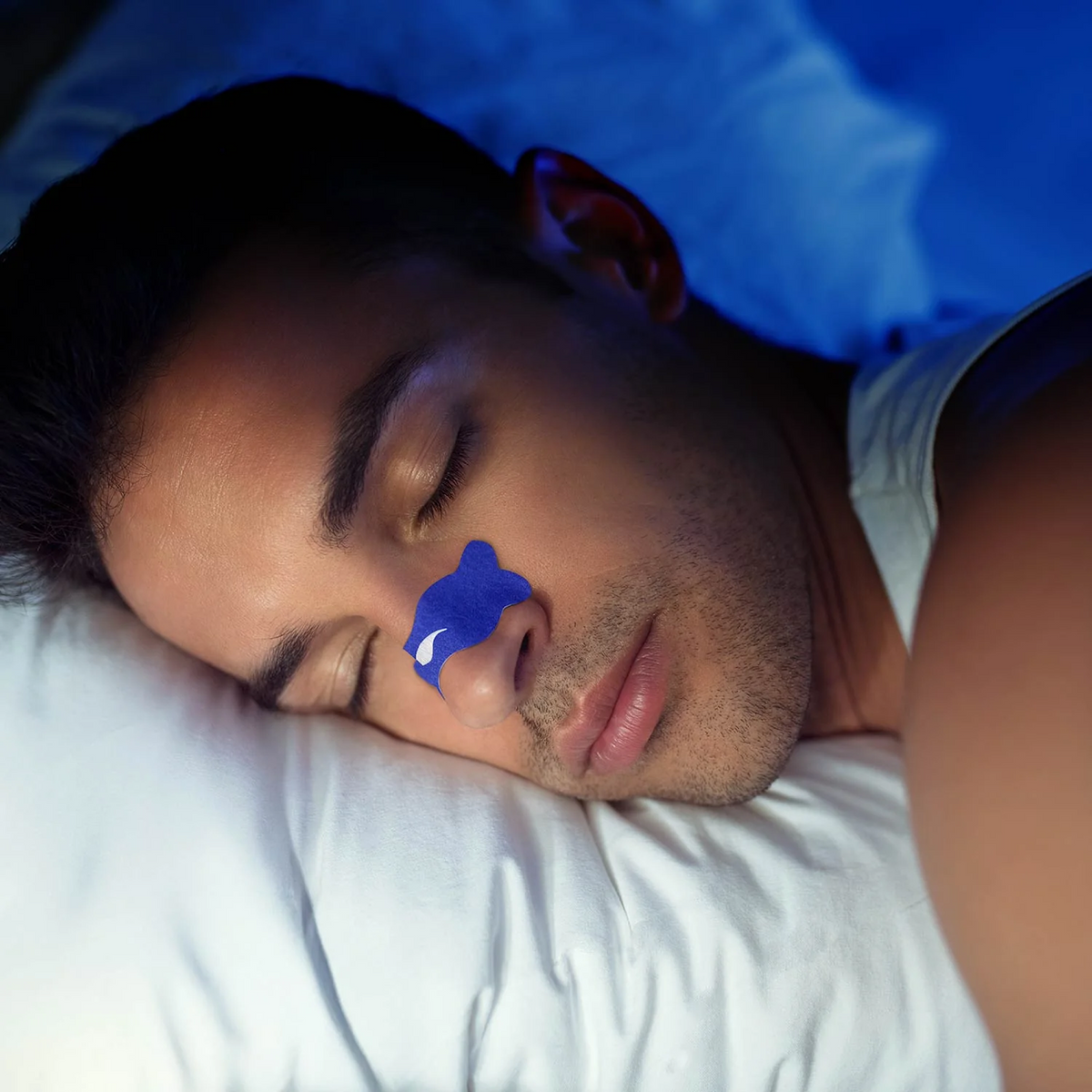 A man sleeping peacefully on a white pillow, wearing a blue Manta Sleep Nasal Strip over his nose, under cool blue night lighting.