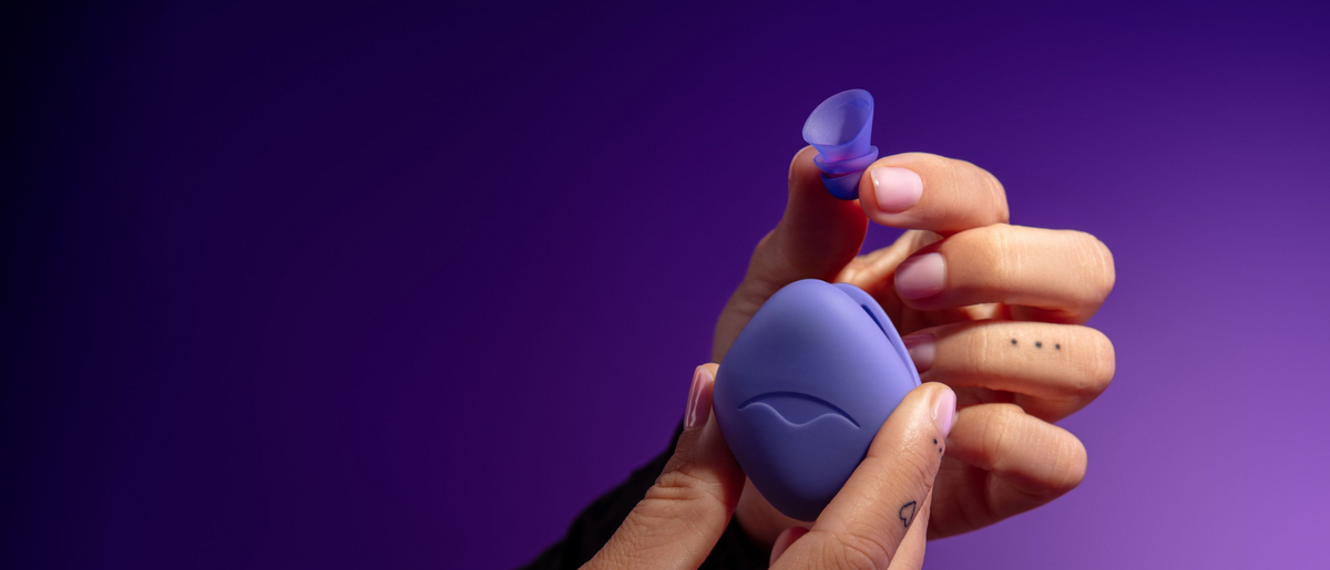 Hands holding a small purple Manta Sleep earplug and its silicone carrying case with Manta Sleep logo against a purple background.