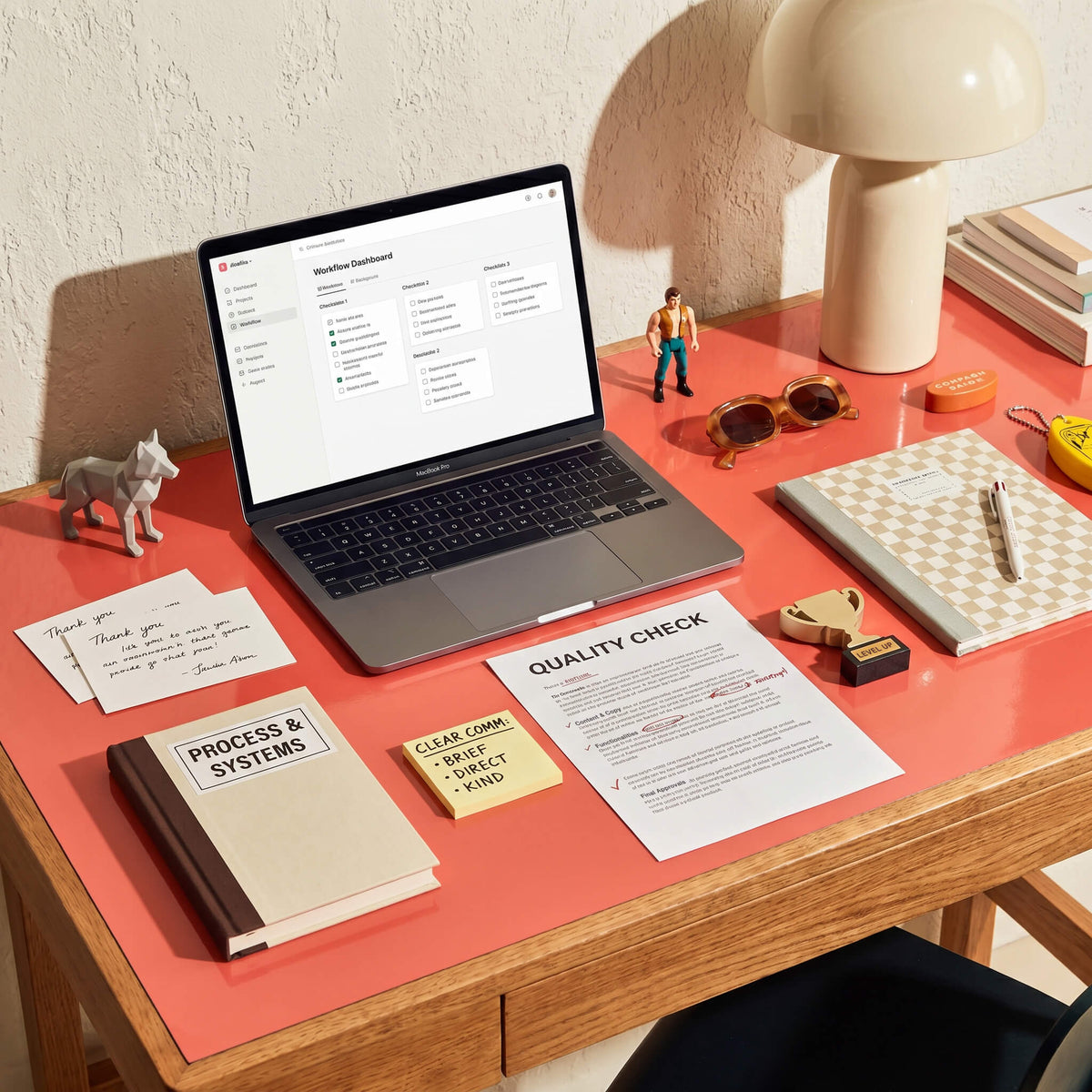 Organized wooden desk with a coral top featuring a laptop showing a workflow dashboard, a quality check document, and a "Process & Systems" book.