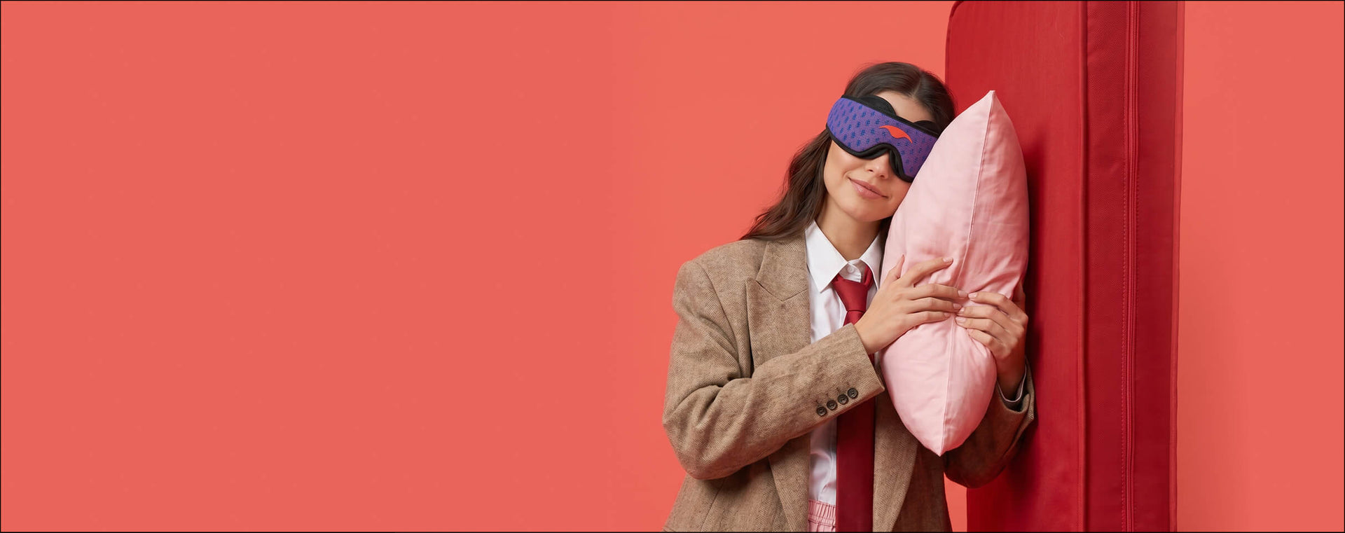 Woman in a business suit and a blue Manta PRO Sleep mask smiling and hugging a pink pillow against a solid coral background.