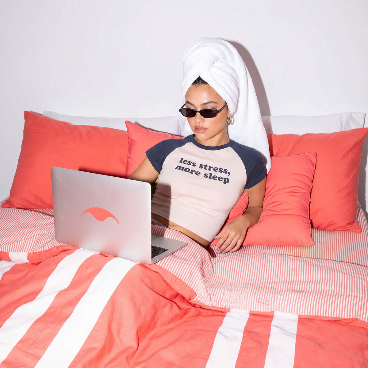 Woman in a "less stress, more sleep" t-shirt, sunglasses, and head towel working on a laptop with a Manta Sleep logo in a pink striped bed.