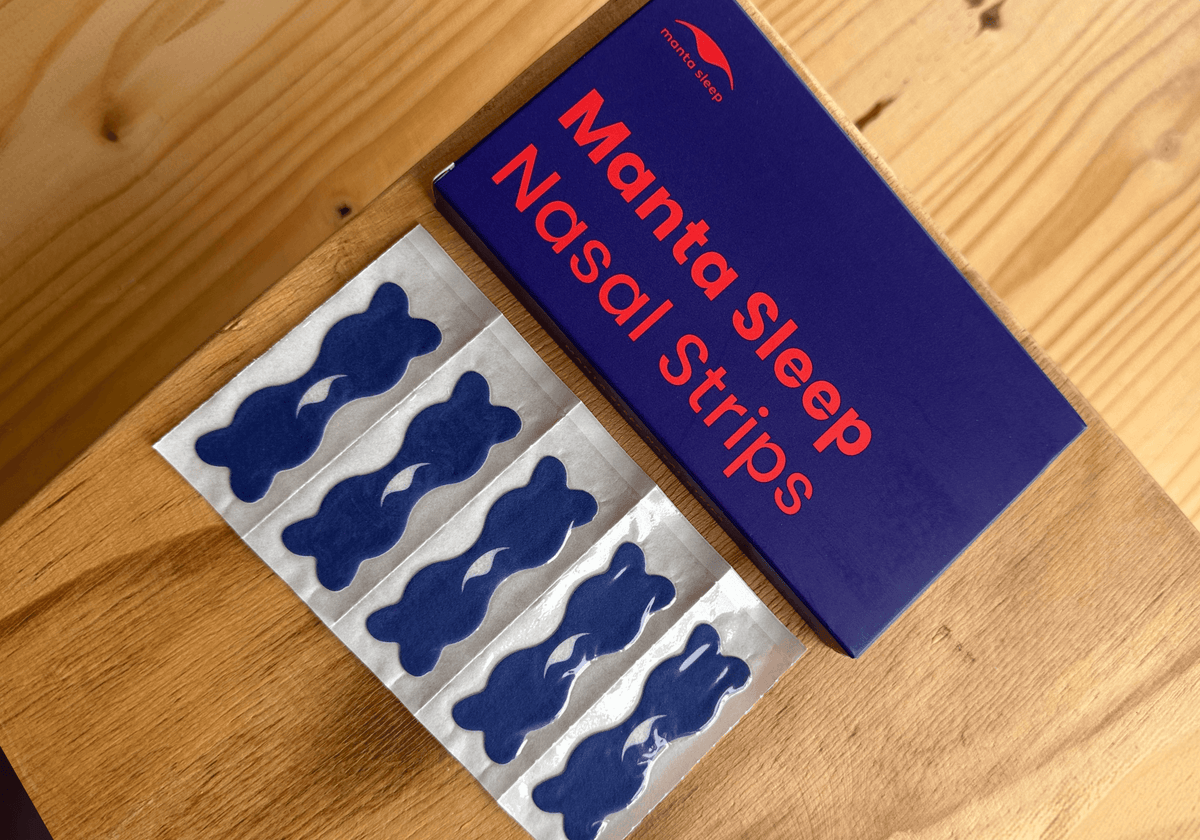 Manta Nasal Strips shown with their dark blue packaging box on a light wood surface, alongside five individual blue nasal strips.