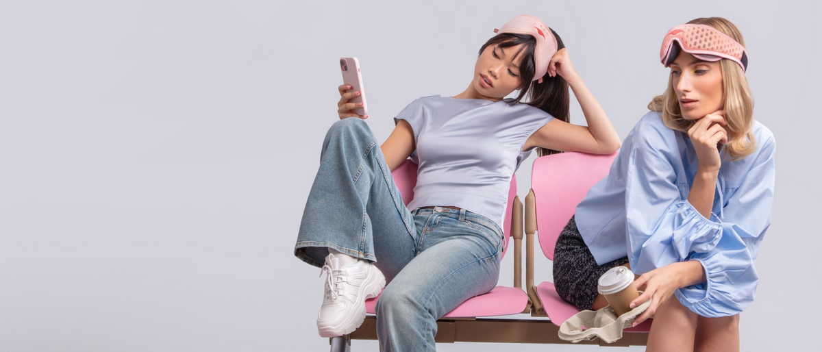 Two women posing in lounge attire on a pink bench against a light gray studio background. Woman on the left wears Pink Spark Manta SOUND Sleep Mask, while woman on the right wears Pink Spark Manta PRO Sleep Mask.