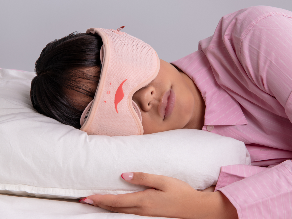A woman sleeping peacefully on a white pillow, wearing a Pink Spark Manta SOUND Sleep Mask.