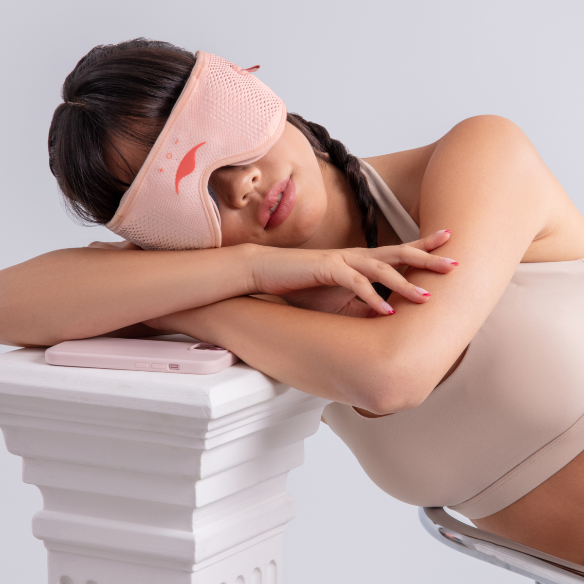 Woman wearing a pink Manta SOUND Mask resting her head on crossed arms over a white pedestal next to a pink phone.
