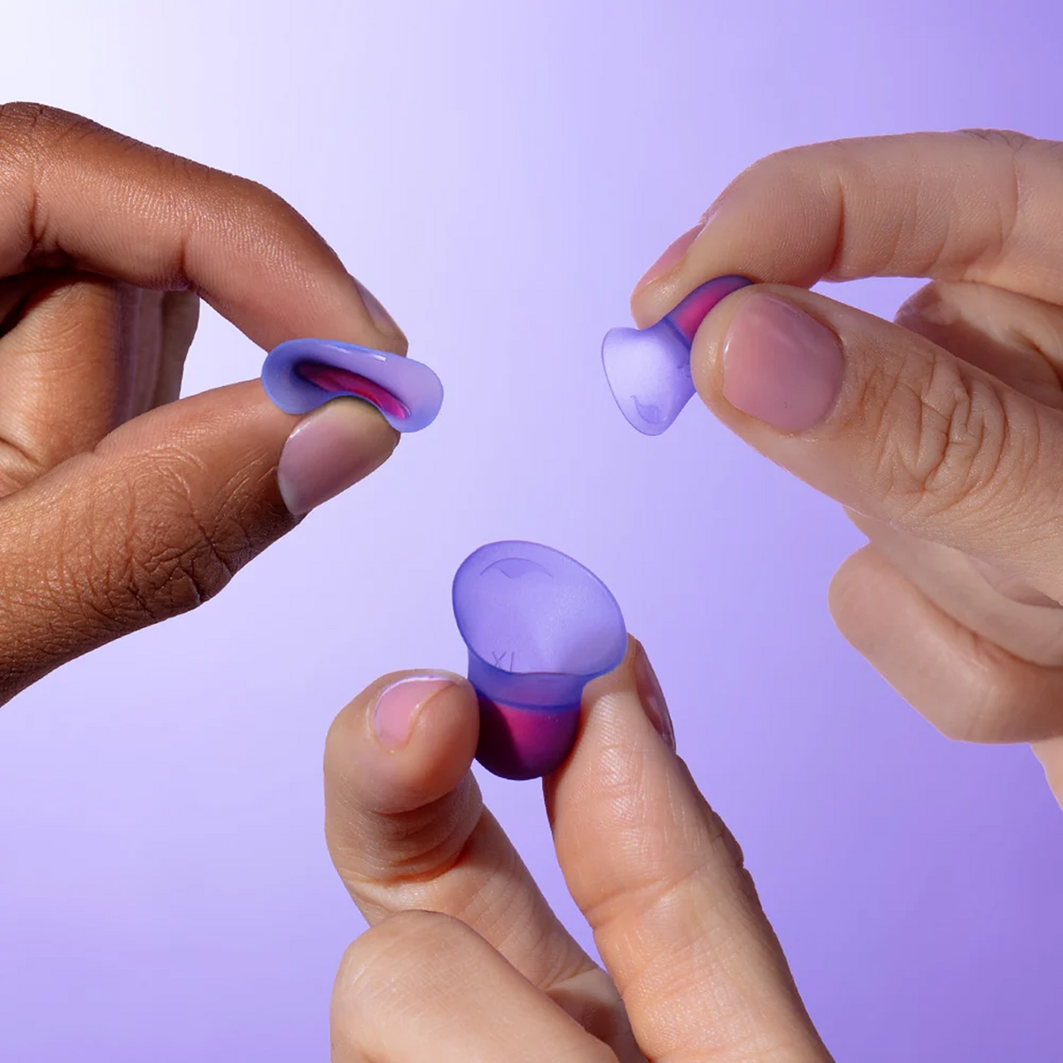 Three hands holding small translucent Manta Sleep Earplugs in purple and coral against a soft lavender background.