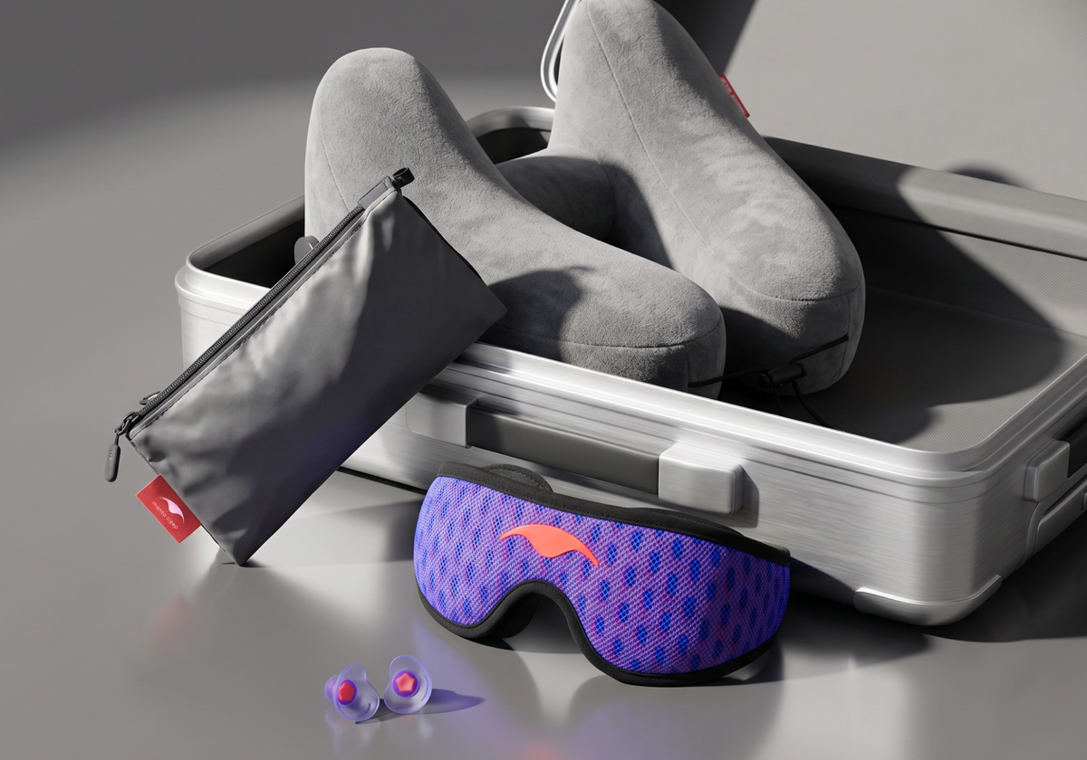 Manta PRO Traveler Bundle in and around an open silver suitcase on a gray studio surface. The set includes a gray Manta Travel Pillow, a gray Manta Travel & Laundry Bag, a blue-purple Manta PRO Sleep Mask, and a pair of Manta Sleep Earplugs.